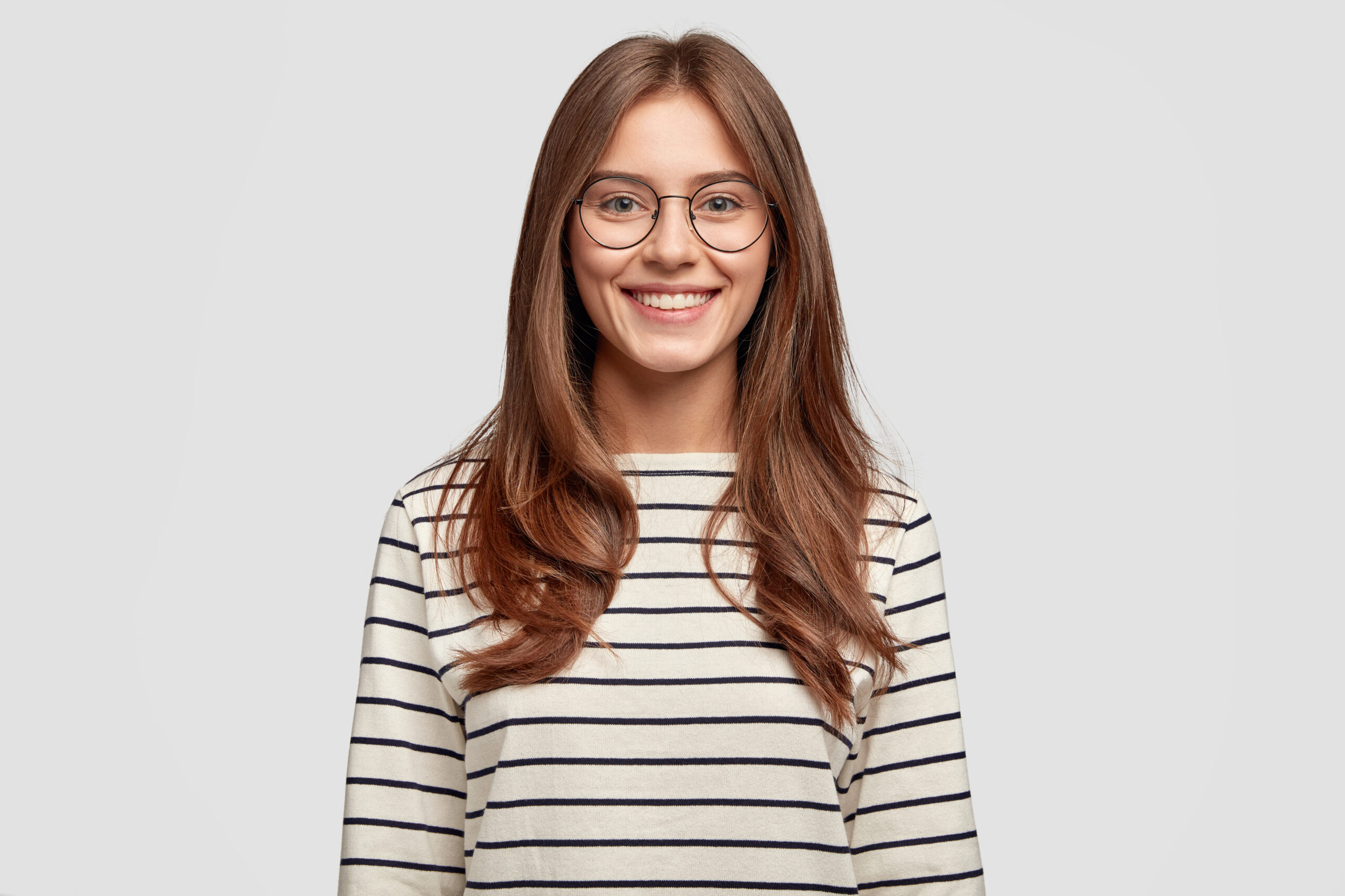 indoor shot of attractive european schoolgirl has toothy smile, dressed in striped sweater, rejoices positive moment in life, wears round spectacles. people, lifestyle and positive emotions concept